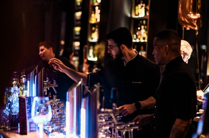 Barkeeper arbeiten hinter einer schwach beleuchteten Bar mit Flaschen und Gläsern und servieren Getränke unter warmem Licht.
