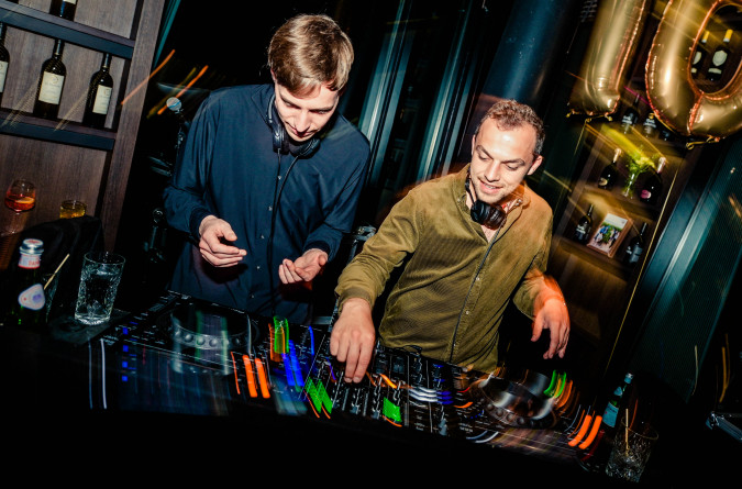 Zwei DJs legen auf einer Party Musik auf, in deren Nähe Getränke stehen, im Hintergrund sind goldene Luftballons mit der Aufschrift „10“ zu sehen, die ein Jubiläum feiern.