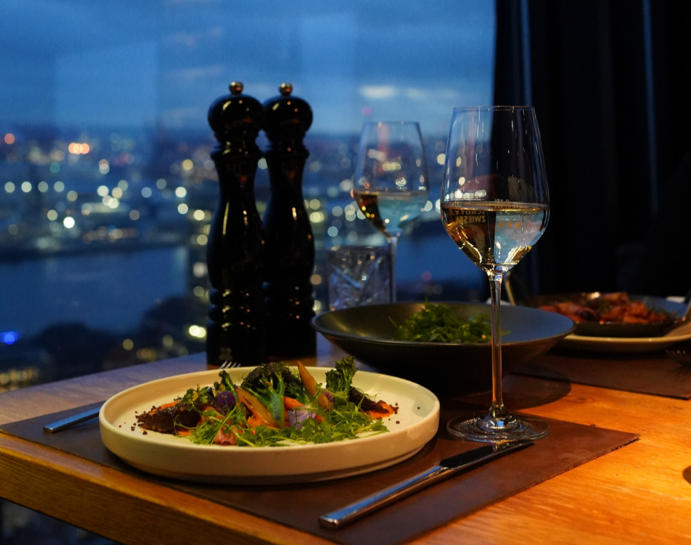 A table set with salad, wine glasses, and pepper grinders overlooks a cityscape at dusk through a window.