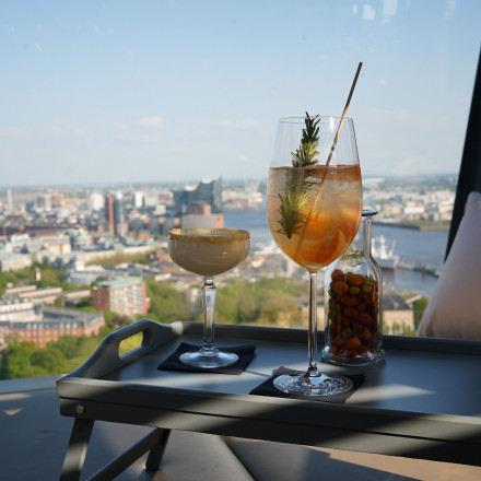 Zwei Cocktails und ein Glas mit Snacks auf einem Tablett, im Hintergrund sind an einem sonnigen Tag eine Stadtlandschaft und ein Fluss zu sehen.