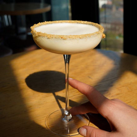 Eine Hand hält ein Cocktailglas mit einem cremigen Getränk und einem mit Krümeln bedeckten Rand, das auf einem sonnenbeschienenen Holztisch neben einem Fenster steht.