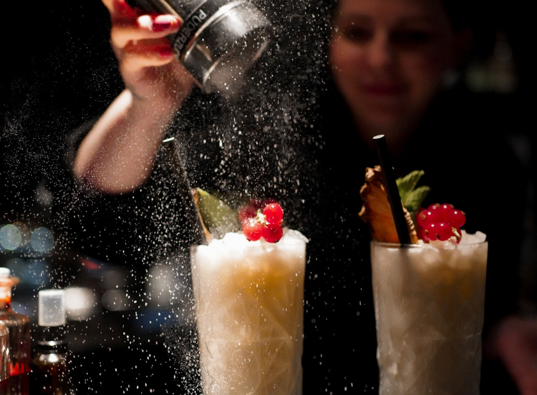 A bartender sprinkles powder over two tropical cocktails garnished with fruit and mint on a bar counter.