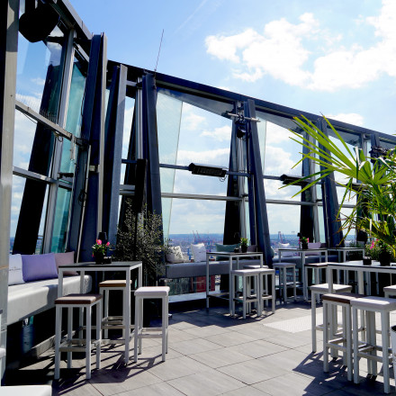 Rooftop bar with tall glass walls, modern tables and stools, potted plants, and a sunny sky in the background.