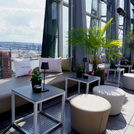 Rooftop lounge with seating, tables, plants, and a view of a busy port.