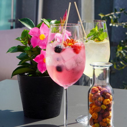Two colorful cocktails and a jar of mixed nuts on a table with a potted pink flowering plant in the background.