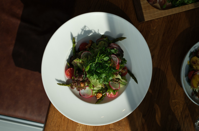 A white plate with a fresh salad of greens, radishes, tomatoes, asparagus, and vinaigrette on a wooden table.