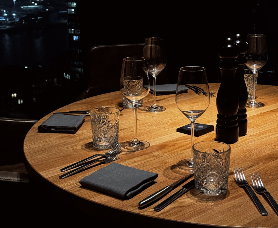 A dimly lit round table set for four with glasses, silverware, and napkins, overlooking a cityscape at night.