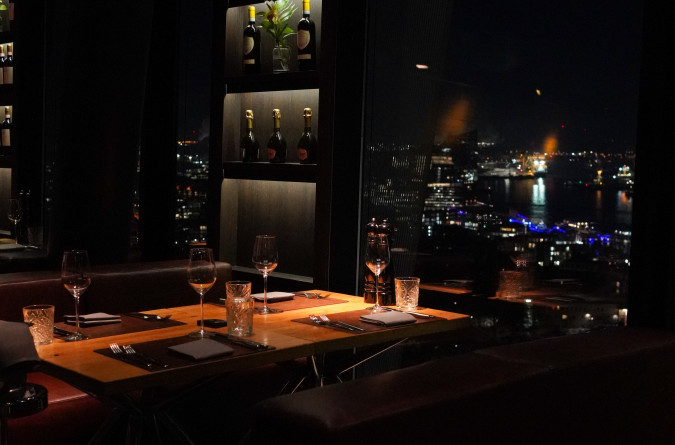 Von einem schwach beleuchteten Restauranttisch am Fenster hat man einen Blick auf die Stadtlandschaft mit funkelnden Lichtern in der Nacht; Weingläser und -flaschen sind zu sehen.