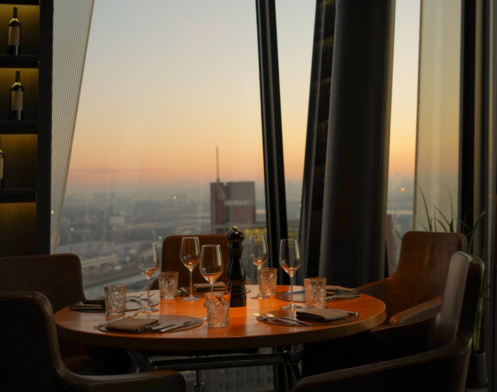 A round table set for four overlooks a cityscape at sunset, with wine glasses and blinds partially open on tall windows.