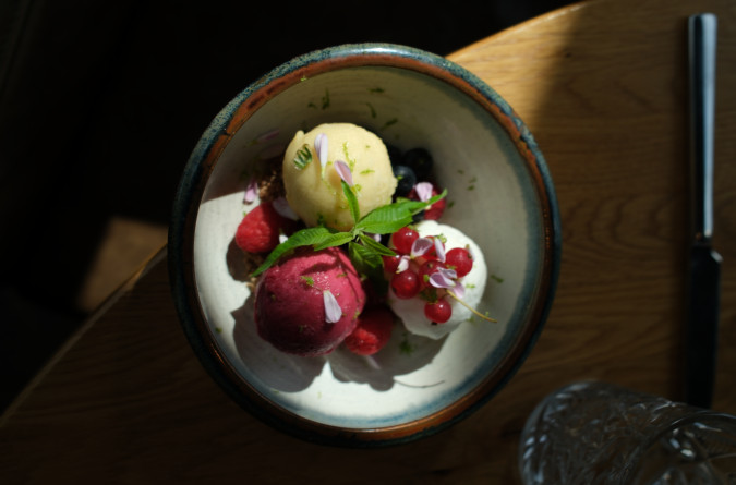 A bowl with three scoops of sorbet, garnished with berries, edible flowers, and a green leaf, on a wooden table.