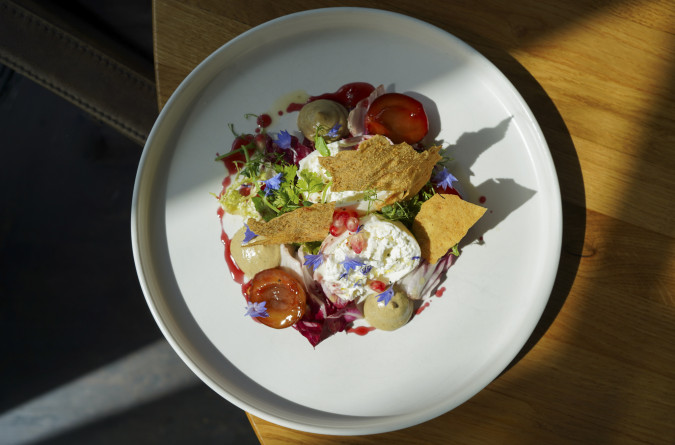 Ein bunter Gourmetsalat mit Käse, Tomaten, Gemüse, essbaren Blumen und knusprigen Crackern auf einem weißen Teller, angerichtet auf einem Holztisch.