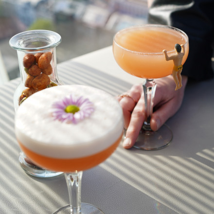 Zwei Cocktails auf einem Tisch, einer mit Schaum und einer Blume garniert, der andere mit einer winzigen Figur; ein Glas Nüsse und im Hintergrund ein Blick auf die Stadt.