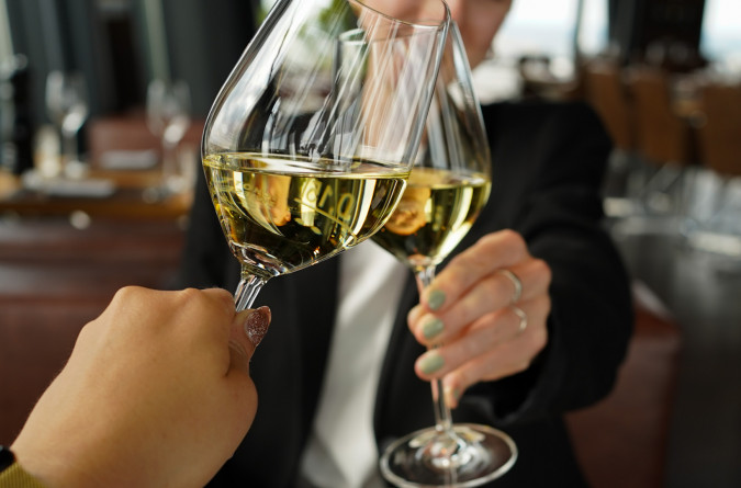Zwei Personen stoßen in einem Restaurant mit Weingläsern an. Vor ihnen stehen Teller mit Essen auf einem Holztisch.