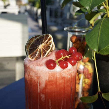 Ein roter, fruchtiger Cocktail mit Schaum, garniert mit roten Beeren und einer getrockneten Limettenscheibe, steht auf einem Tisch neben einer Pflanze.