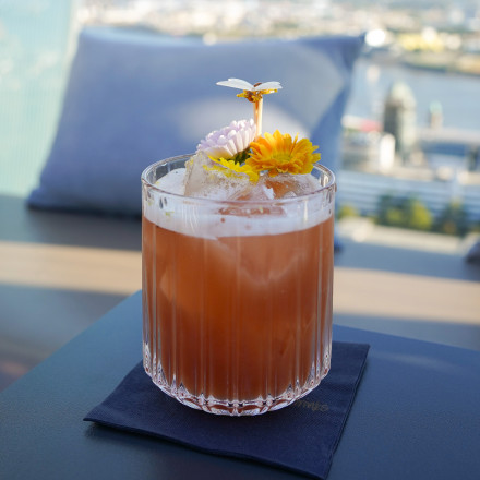 Ein rosa Cocktail mit Eis und essbaren Blüten wird in einem Glas auf einer Serviette serviert, im Hintergrund ist eine verschwommene Stadtlandschaft zu sehen.