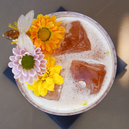 Ein Cocktail mit Eiswürfeln, Schaum, bunten Blumen und einer dekorativen Biene obendrauf, von oben auf einer dunklen Serviette gesehen.