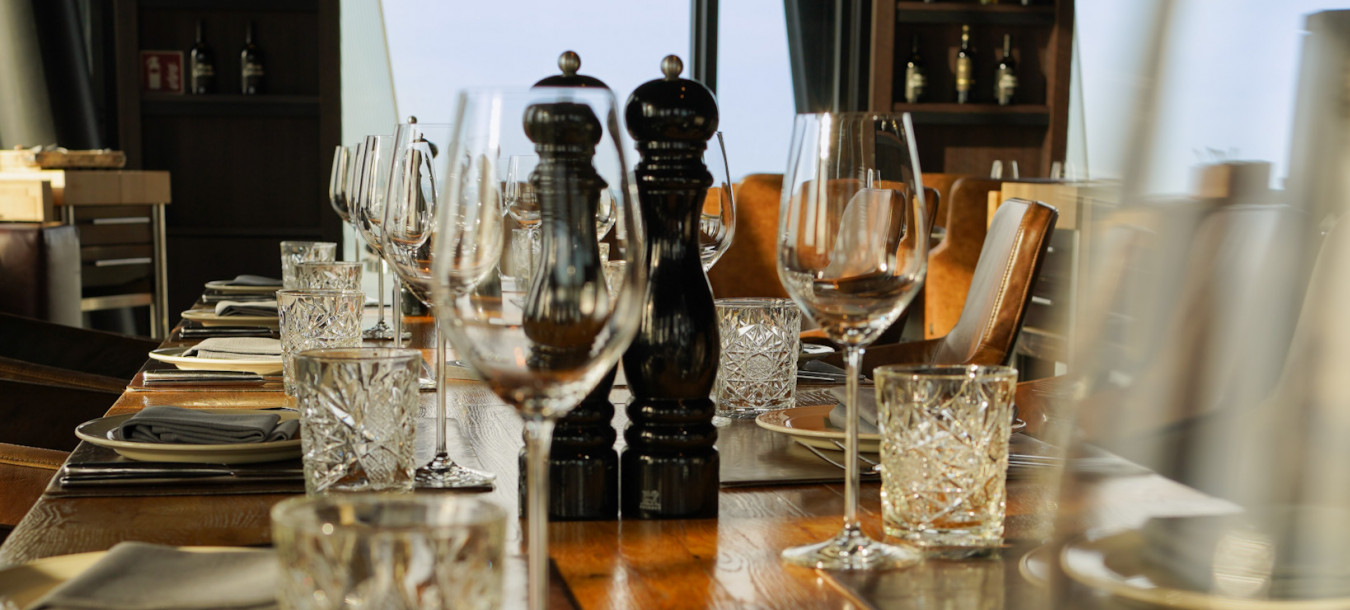 A set dining table in a modern restaurant with crystal glasses, plates, and pepper mills, sunlight streaming through large windows.