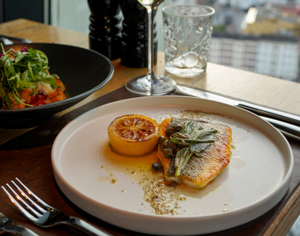 A plate with grilled fish and lemon, salad bowl, wine glass, pepper grinders, and city view from a high-rise window.