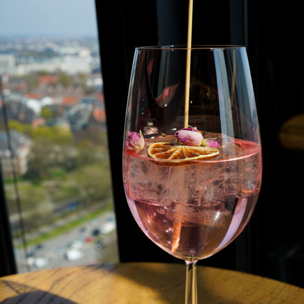 Ein Glas rosa Cocktail mit Eis, getrockneten Zitrusscheiben und Rosenblättern auf einem Holztisch neben einem sonnigen Fenster mit Blick auf die Stadt.