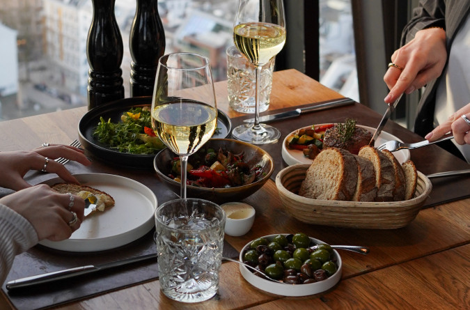Two people dining by a window with wine, bread, olives, and vegetables on the table, overlooking a cityscape below.