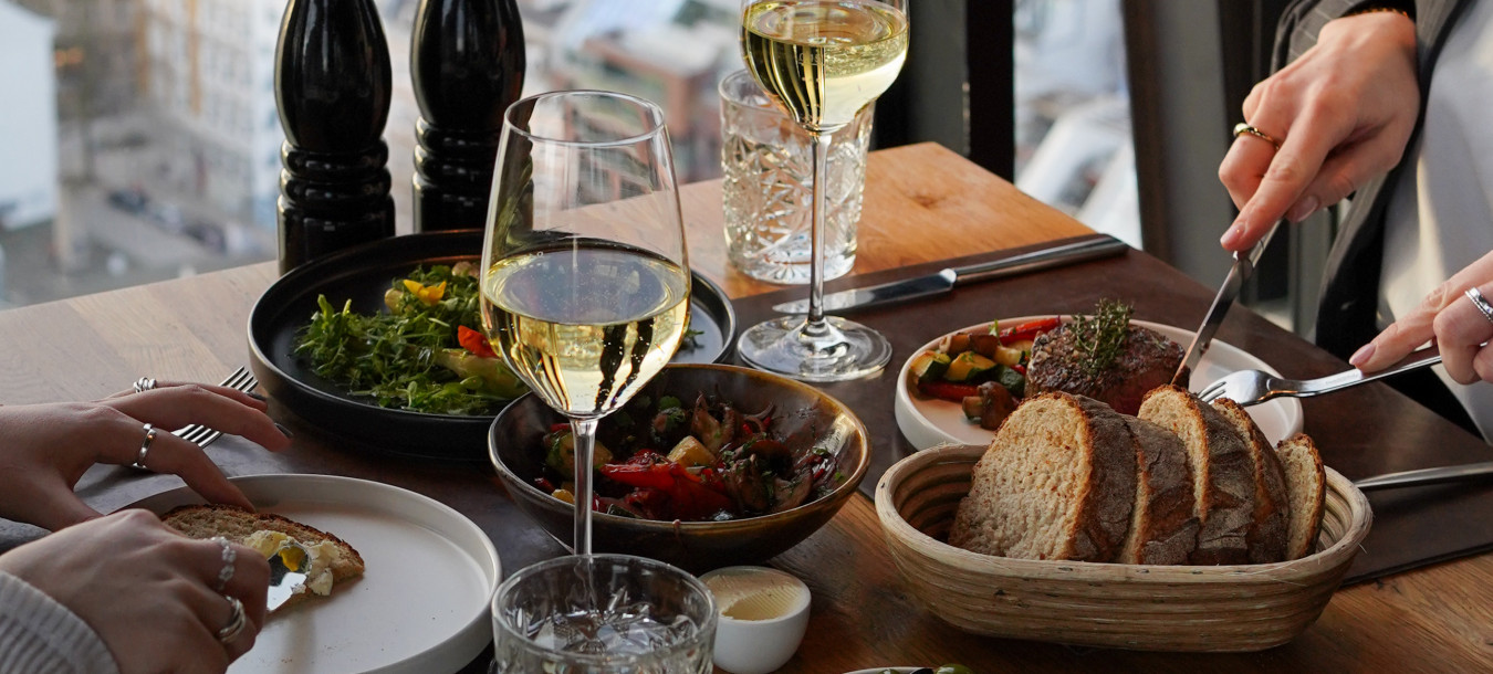Two people dining at a restaurant with wine, bread, olives, and dishes on a table by a window overlooking a cityscape.