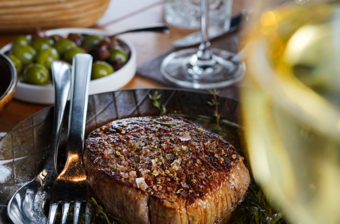 Cooked steak with herbs and garlic on a plate, beside utensils, olives, bread, and a glass of white wine on a table.