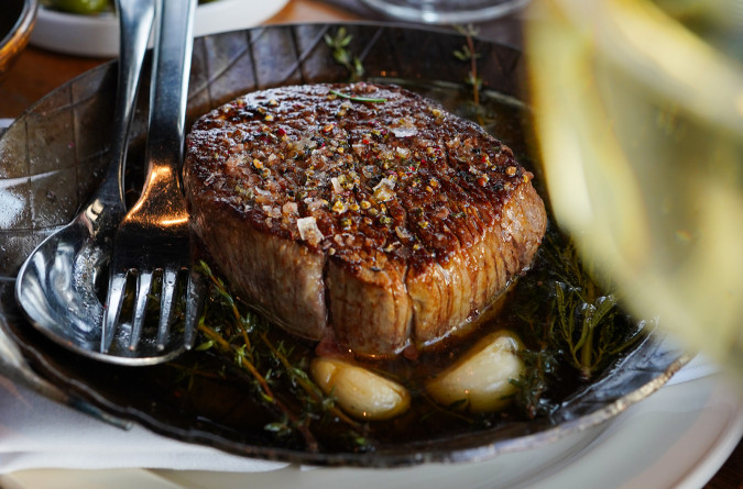 Ein gegrilltes Steak auf einem Teller mit Kräutern und Knoblauch, daneben Besteck, im Hintergrund Wein, Oliven und Brot.