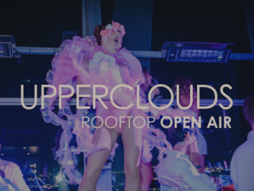 Ein Künstler in einem rosafarbenen Kostüm unterhält die Gäste einer Dachveranstaltung, wobei der Text UPPERCLOUDS ROOFTOP OPEN AIR überlagert wird.