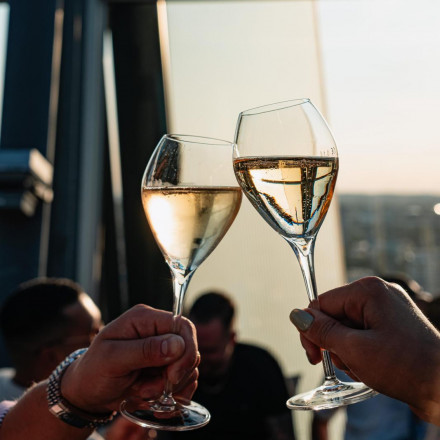 Zwei Hände stoßen mit Weingläsern an, im unscharfen Hintergrund sind Menschen und Sonnenlicht zu sehen.