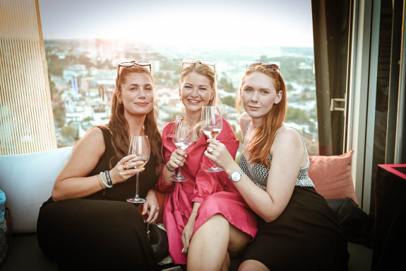 Drei Frauen sitzen dicht beieinander, lächeln und halten Weingläser, im Hintergrund ist ein Blick auf die Stadt zu sehen.